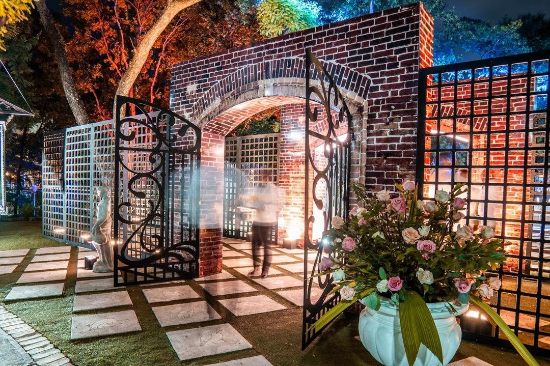 Brick archway with wrought-iron gates, lit at night, roses in the foreground